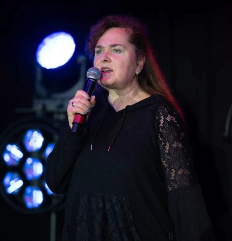 Frau singt mit Mikrofon auf der Bühne, beleuchtet von Scheinwerfern.