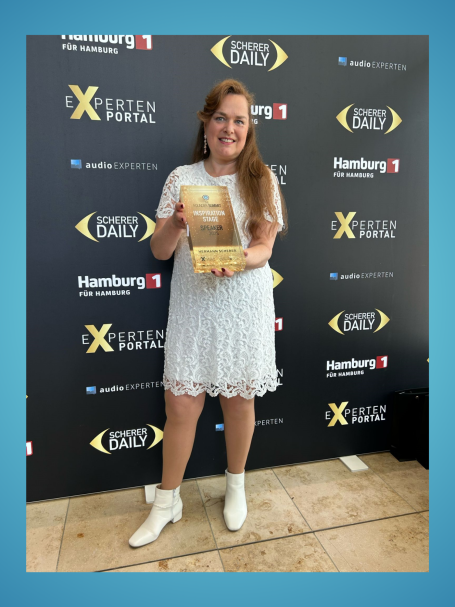 Frau in weißem Kleid hält einen Preis vor einer schwarzen Wand mit Logos.